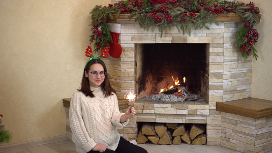 A young pregnant woman in glasses sits by the fireplace with a sparkler in her hand. Christmas mood. 4k