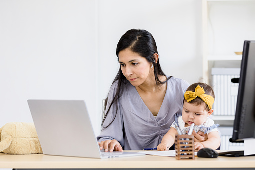 Working from her home office, the mid adult female customer service representative holds her baby daughter as uses her laptop to help a customer.