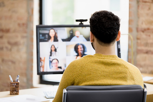 The focus of the photo is on the back of the unrecognizable man as he attends a virtual staff meeting.