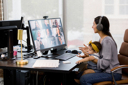 The mid adult mother, holding her baby, uses a co-working space to video conference with her co-workers.