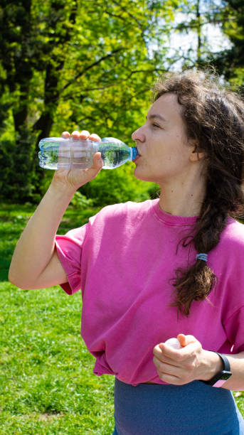 istock Young woman drinking clear cool mineral water from plastic bottle after training, standing in green park garden outdoors. Maintaining water balance in hot summer day. Thirst quencher. Vertical 1396725020