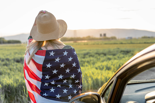 4th of July. Fourth of July. American woman with the national American flag against beautiful landmark. Independence Day. Beautiful sunset light.