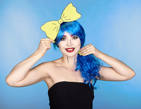 Portrait of young woman in comic pop art make-up style. Female in blue wig on blue background. Girl with yellow bow-tie in hands