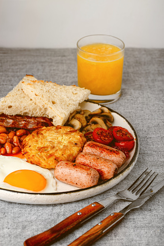 English breakfast with fried eggs, hash brown, sausages, bacon, beans, toasts, mushrooms, tomato and orange fresh juice.