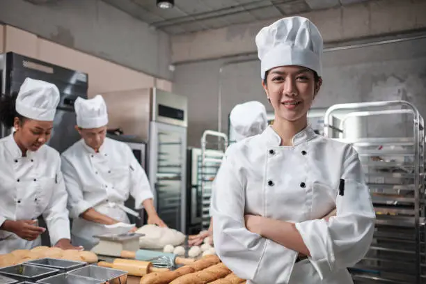 Young beautiful Asian female chef in white cooking uniform looks at camera, arms crossed and cheerful smile with food professional occupation, commercial pastry culinary jobs in a restaurant kitchen.