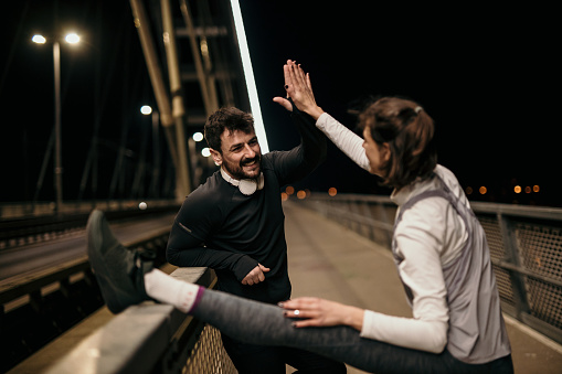Spontaneous photo of two, fitness man, bearded, in a black active wear and a white headphones, and woman, a brunet, wearing a gray, white and purple colors of active wear, giving each other a high five after the training session outdoors done, at night. Radiating happy