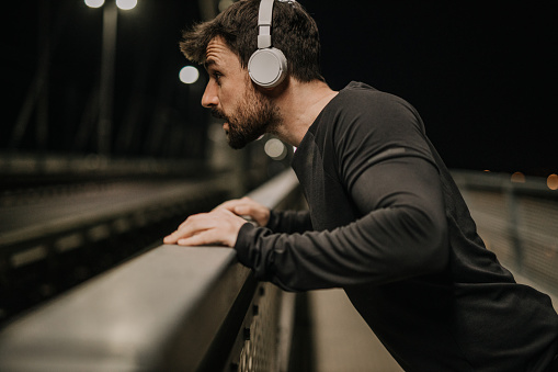 A photo of a handsome bearded man, the athlete, with headphones on head doing the push ups while leaning on the fence of the bridge, a night workout in the city. He's in his black active wear