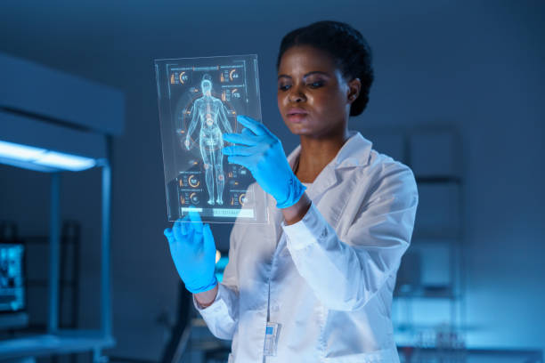 A young concentrated African - American scientist in a lab coat and protective gloves works on a small HUD or graphic display with the image of a human body stock photo