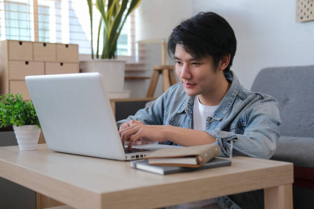 istock Smiling handsome asian man sitting on floor in living room and using laptop computer. 1396806882