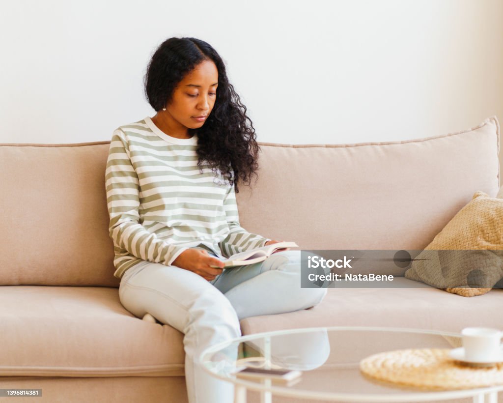 Beautiful African-American female sitting on couch and reading book. Spending time at home alone African-American woman in casual comfy clothes reading book while sitting on sofa in cozy living room. Home interior. Concept of maintaining emotional wellbeing. Having micro-moment and enjoying life Digital Detox Stock Photo