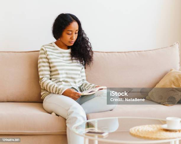 Beautiful Africanamerican Female Sitting On Couch And Reading Book Spending Time At Home Alone Stock Photo - Download Image Now