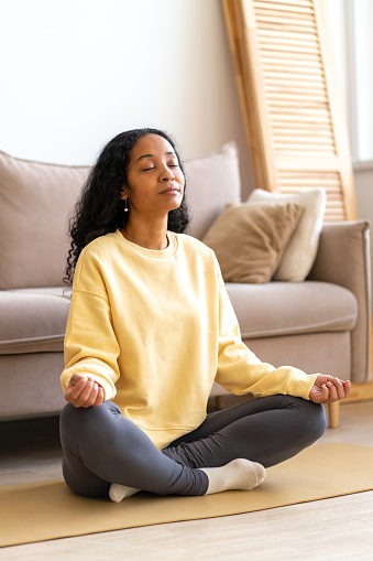 African-American woman in sportswear bare feet sitting in yoga lotus position on mat in living room while meditating and doing breathing therapy. Concept of mindfulness and stress relief
