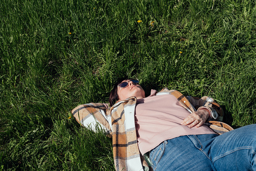 Woman in sunglasses lies on the fresh grass covering her face from the sun with her hand. Carefree time, freedom and tranquility. Copy space