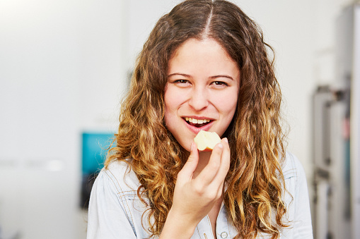 Smiling, happy young woman eats a piece of fruit, with copy space on the kitchen wall.
