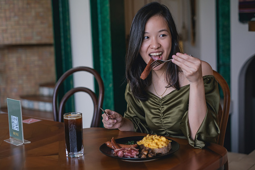 Candid shot of a beautiful young Southeast Asian single woman eating continental breakfast in a restaurant while traveling. She's having an iced Americano with scrambled egg and bacon on top of a toast.