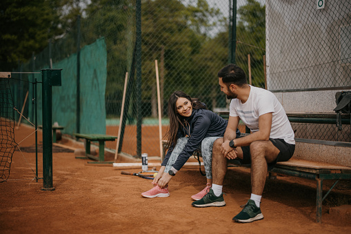 Young couple of tennis players sitting on bench on tennis court and preparing for the match, woman is tying shoelace