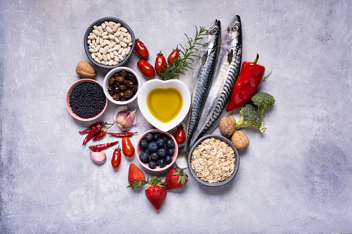 Front view of many fish oil capsules spilling out from the bottle surrounded by an assortment of food rich in omega-3 such as salmon, flax seeds, broccoli, sardines, spinach, olives and olive oil.