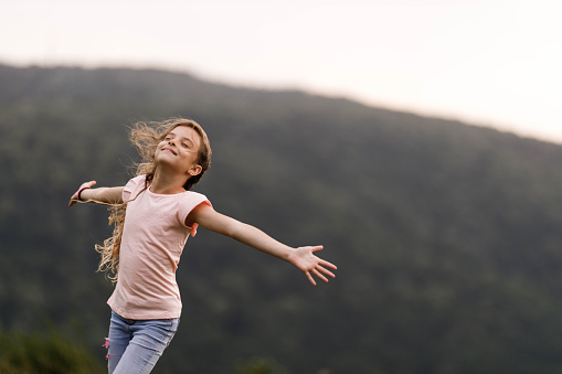 Happy little girl having fun with her arms outstretched and eyes closed during springtime in nature. Copy space.
