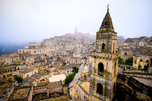 A suggestive view with the fog that envelops the splendid maze of alleys and stone houses of the old town of Matera, in southern Italy, known worldwide as the 'Sassi di Matera' (Matera's Stones). In the foreground on the right the bell tower of the rupestrian church of San Pietro Barisano, originally built in the 12th century. The ancient city of Matera, in the region of Basilicata, is one of the oldest urban settlements in the world, with a human presence that dates back to more than 9,000 years ago, in the Paleolithic period. The Matera settlement stands on two rocky limestone hills called 'Sassi' (Sasso Caveoso and Sasso Barisano), where the first human communities lived in the caves of the area. The rock cavities have served over the centuries as a primitive dwelling, foundations and material for the construction of houses, roads and beautiful churches, making Matera a unique city in the world. In 1993 the Sassi of Matera were declared a World Heritage Site by Unesco. Super wide angle image in High Definition format.