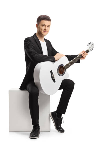 istock Young man in a black suit with an acoustic guitar sitting on a white cube 1396831751