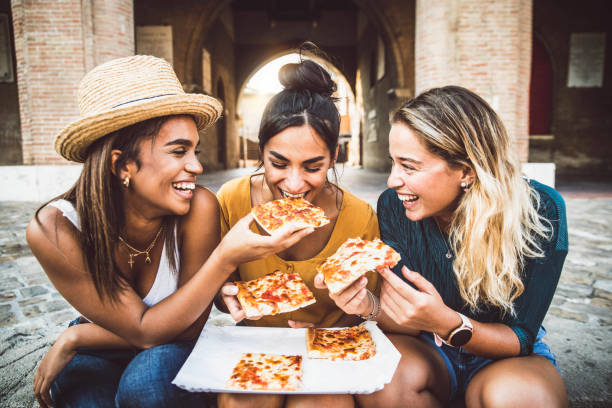 tre giovani amiche che mangiano la pizza sedute fuori - donne felici che gustano il cibo di strada in città - cultura gastronomica italiana e concetto di vacanze europee - teenager city life laughing group of people foto e immagini stock