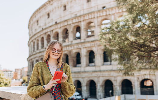 Portrait of a cheerful happy woman on background of Coliseum in Rome, Italy. Beautiful smiling female in dress using smartphone outdoors. Traveler