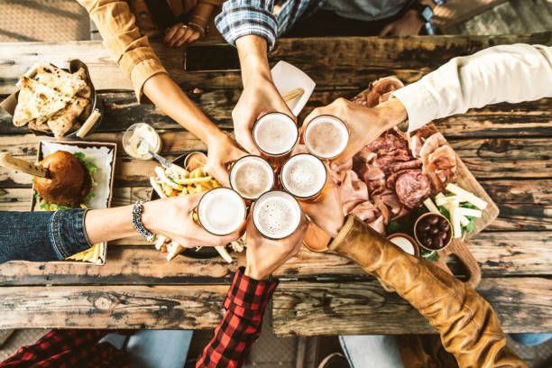 amici che rallegrano i bicchieri di birra sul tavolo di legno coperto di cibo delizioso - vista dall'alto delle persone che cenano al bar ristorante - concetto di stile di vita di cibo e bevande - pub food foto e immagini stock