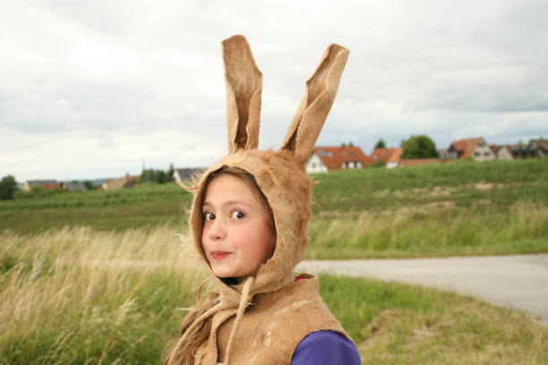 süßes osterhasenmädchen mit den schönen feldern und dem dorf im hintergrund. mädchen mit braunem gefilztem hasenhut. hasenohren filzhut. filzen. gefilzte kunst. gefilzter hut. handgefertigter hut - easter easter bunny fun humor stock-fotos und bilder