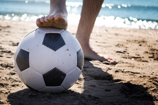 MAN ON THE BEACH HOLDING A SOCCER BALL WITH ONE FOOT OVER THE SAND AND THE SEA AND THE BLUE SKY IN THE BACKGROUND ON A SUNNY SUMMER DAY