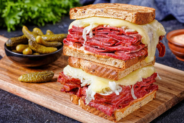 reuben sandwich with corned beef, top view close-up of reuben sandwich with rye bread thinly sliced corned beef, sauerkraut, russian  dressing and melted cheese on board with pickles reuben sandwich stock pictures, royalty-free photos & images