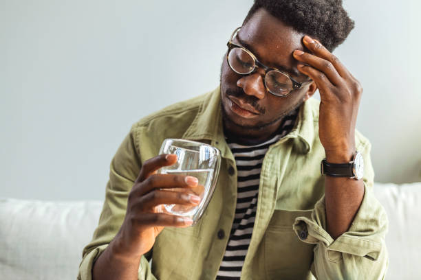 Young man suffering from strong headache Young man suffering from strong headache or migraine sitting with glass of water in the living room, millennial guy feeling intoxication and pain touching aching head, morning after hangover concept substance abuse stock pictures, royalty-free photos & images