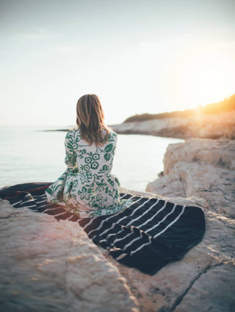 Rear view of lonely young woman sitting on rocky shore.