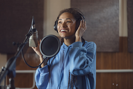 African American female singer records a new song or actress voices text, rehearses with enjoyment and emotions in soundproof room of music studio.