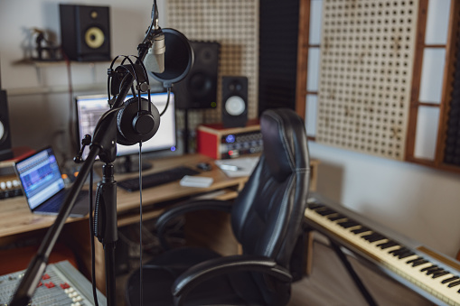 Focus on microphone and headphones in the blurred sound engineer office for broadcasting and music rehearsal space with modern mixing board, software, laptops and synthesizer. Sound recording studio