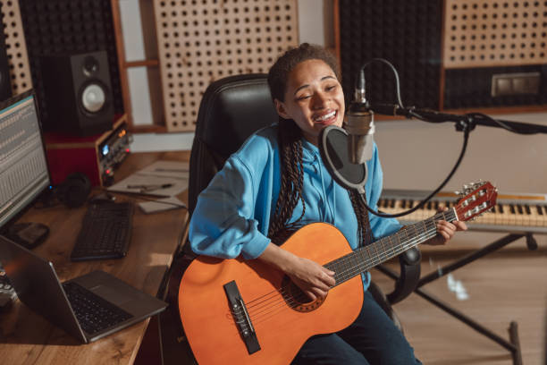 istock Cute young African musician singing into microphone while performing new song and playing guitar in the recording studio 1396841792