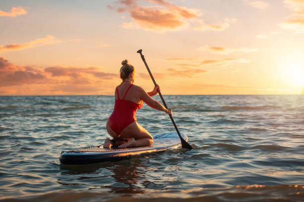 plus sized beautiful caucasian woman in red swimsuit swimming sitting on a sup board. sunset on the background. back view. the concept of sports and summer vacation - women paddleboard bikini surfing imagens e fotografias de stock