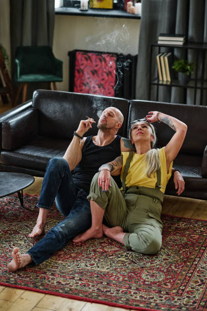Smoking Cannabis In Living Room Vertical high angle view of modern mature Caucasian couple relaxing on floor in loft living room smoking marijuana marijuana tattoo stock pictures, royalty-free photos & images