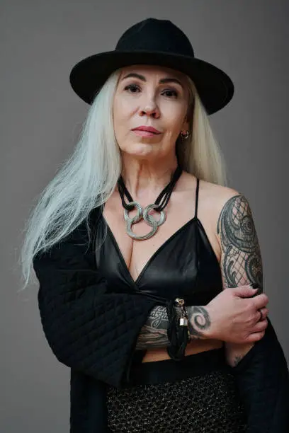 Vertical medium portrait of sensual mature Caucasian woman with long gray hair and tattooes on arms posing on camera