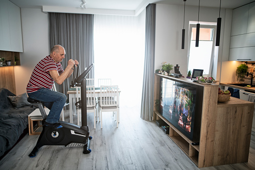 Senior man is spending time at home. He  is exercising using a exercise bike and watching family hiking movie on TV. 
Canon R5