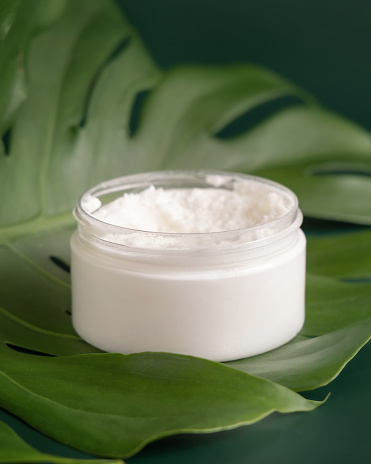 Opened White jar on monstera leaf on green table close up. Brand packaging mockup. Skincare beauty product, cream or butter. Making homemade herbal cosmetics from natural ingredients