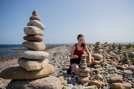 Wide shot selective focus shot of a young woman building a pebble stack while at a beach at Holy Island in the North East of England in summer.
