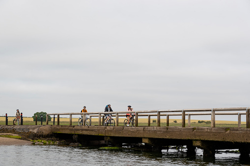 Wide angle shot of a group of mixed ethnic teens on a  bike ride together at Holy Island in the North East of England in summer. They are riding over a bridge by the ocean.