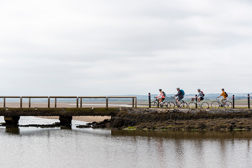 Distant wide angle side view shot of a group of mixed ethnic teens on a  bike ride together at Holy Island in the North East of England in summer. They are riding over a bridge by the ocean.