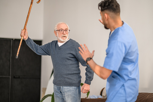 Irritated gray-haired old man brandishing the walking stick at his in-home caregiver standing before him