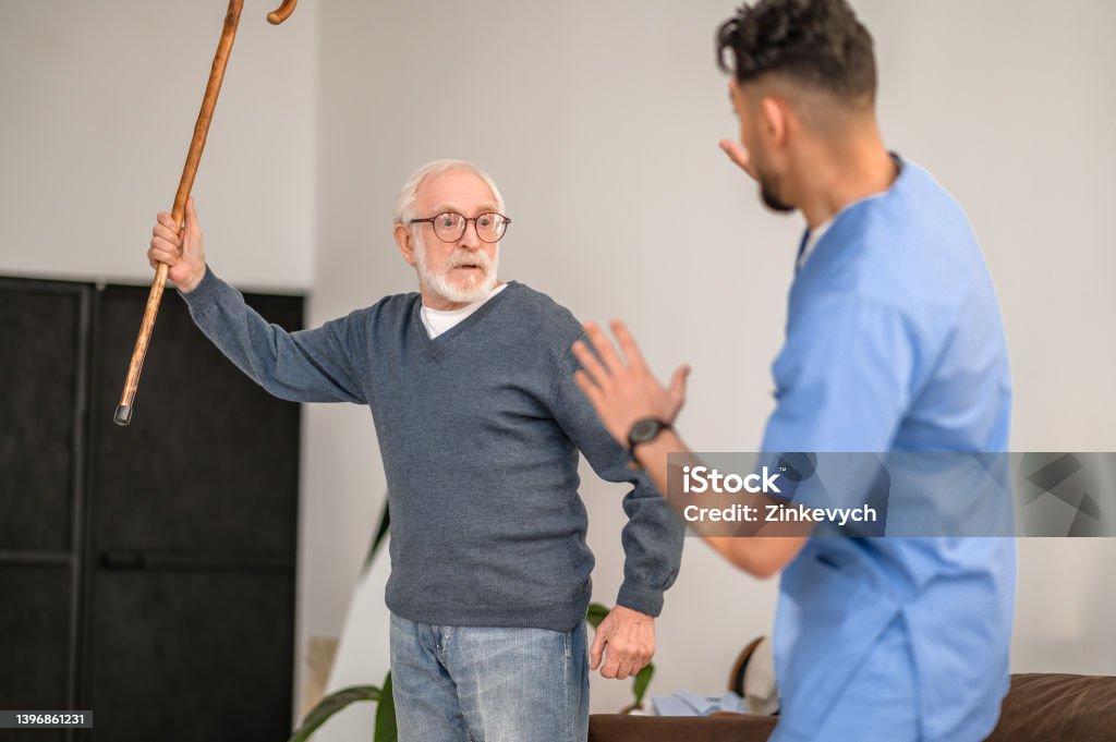 Exasperated pensioner threatening his caretaker with his cane Irritated gray-haired old man brandishing the walking stick at his in-home caregiver standing before him Anger Stock Photo