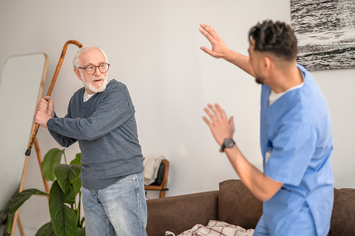 Angry aged person shaking his cane at the young in-home male nurse standing before him