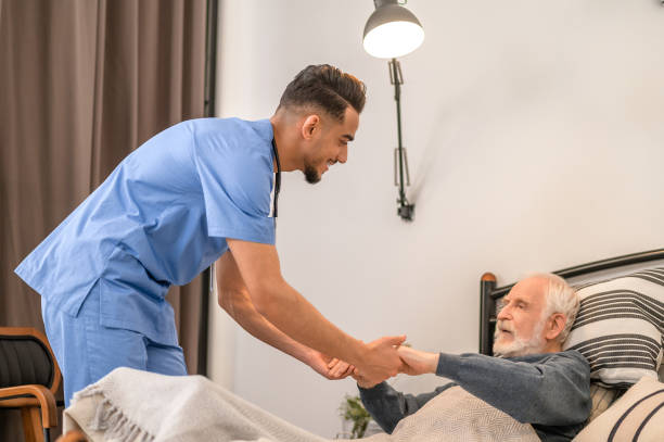 Smiling medical worker lifting the old man from the bed stock photo
