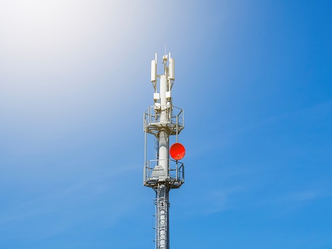 A 5G telecommunications tower on the background of a clear blue sky without clouds. A gray cellular antenna with a red satellite dish.