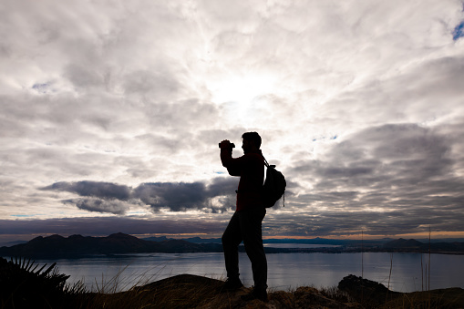 Silhouette of a young Hiker looking into the distance with binoculars in front of the landscape on the north coast of Mallorca with the bay of Pollensa in the background. Color editing. Part of a series.