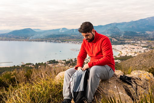 Happy dressed with a red sweatshirt sitting on a rock in the mountains looking with his backpack on the beautiful north coast with the bay of Pollensa in the background of the island Majorca. Color editing. Part of a series.